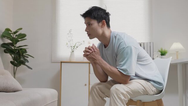 An Asian man, looking stressed about household finances, slumps down in his living room, showing signs of worry.