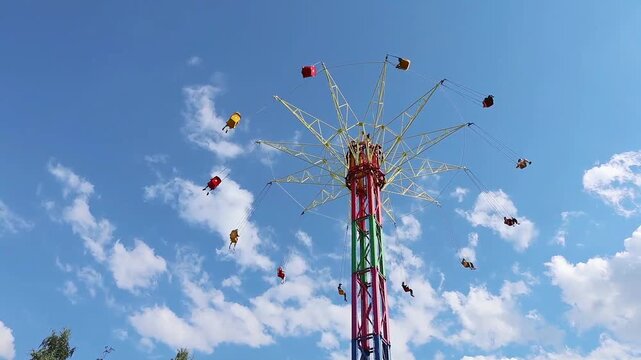 Colorful chain swing amusement ride with riders suspended in mid-air against a bright blue sky with fluffy white clouds, vibrant steel structure in red, green, yellow, and magenta tones