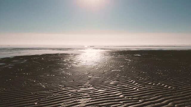Horizontal 4K 24fps low altitude aerial drone video flying forward over wet sand ripple patterns on a beach during sunset in Grado, Italy, with bright sun reflecting on the calm sea