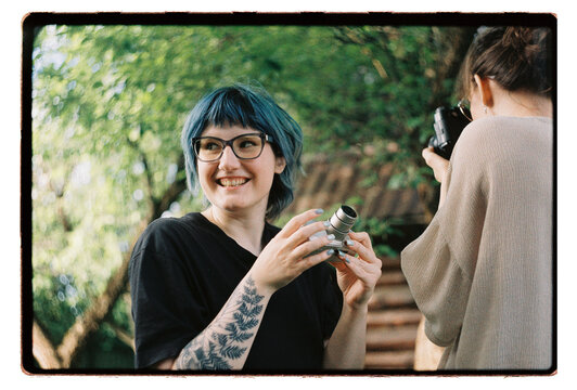A woman with blue hair laughs while holding a camera lens outdoors