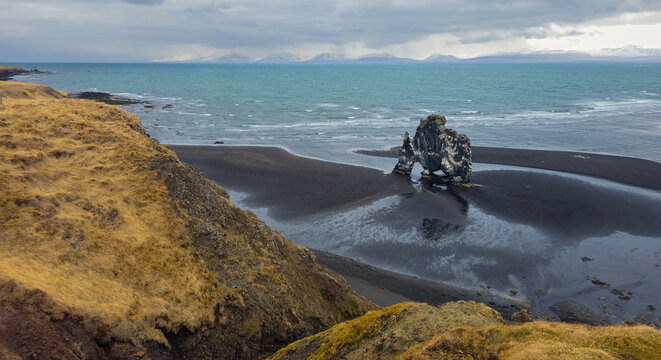 Aerial view of the Hvitserkur basalt sea stack on a black sand beach, with snow-capped mountains in the distant background under a cloudy sky.