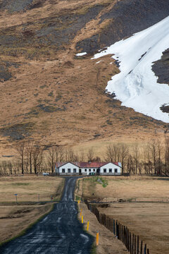 Winding country road leading to a remote farmhouse under a massive mountain with melting snow in Iceland.