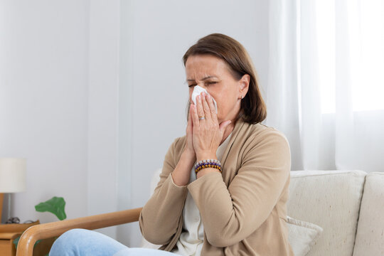 Mature woman sneezing with tissue sitting on a couch feeling unwell in living room