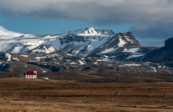 Horizontal the historic Ingjaldsholskirkja Ingjaldsholl Church in western Iceland, with majestic snow-dusted mountains rising dramatically under a cloudy blue sky.