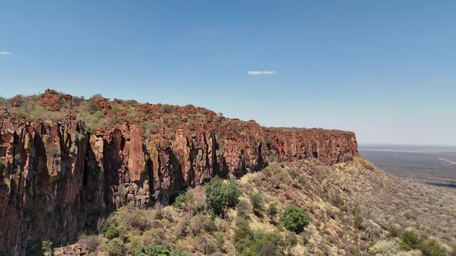 Cinematic aerial footage of a massive red sandstone escarpment towering over the dry, flat savanna plains in Namibia, Africa. Epic geological landscape.