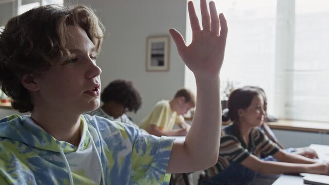 Caucasian teenage boy raising hand to answer question during high school lesson with classmates in background. Clip for educational marketing and school promotional videos