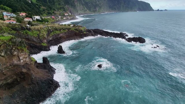 Drone aerial view of rugged coastal cliffs with green vegetation, colorful houses, and turquoise ocean waves crashing against sea stacks in Madeira Portugal.