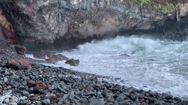 Turbulent waves crash against a rocky cobble beach and layered cliff face on the coastline of Madeira Portugal during daytime.