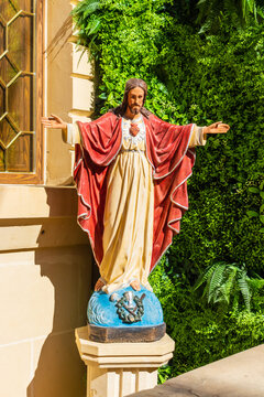 Painted Jesus Statue With Sacred Heart Standing on Pedestal in Garden Near Church Window