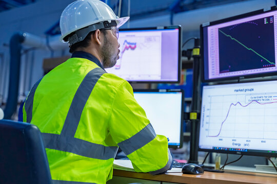 Industrial engineer or technician using a calculator and writing on a notebook while monitoring data on computer screens in a factory control room.