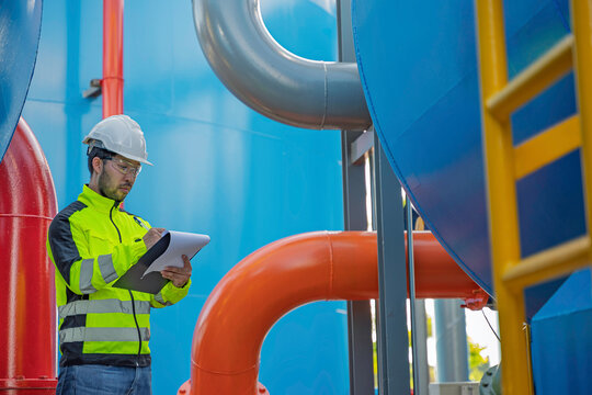 An Asian engineer points at a complex industrial pipeline system, checking water treatment valves and auditing machinery infrastructure for facility maintenance and factory safety control.