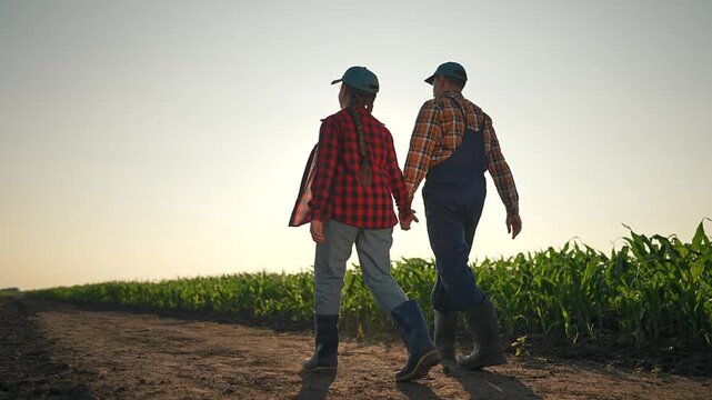 Man in farmer outfit and woman with braid walk field. Couple hold hand by corn at sunset. Path shows fresh boot prints. Corn field lines side. Farmer leads woman down rural path sharing sunset view.