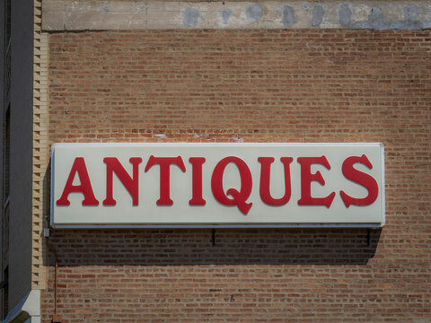 Large white commercial sign with bold red lettering reading Antiques mounted on an old weathered red brick building facade in sunlight