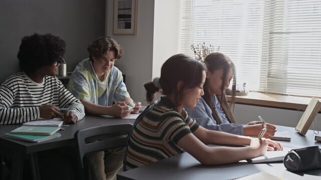 Diverse teenagers sitting at desks in classroom, passing secret sticky note and whispering during lesson. Footage for high school marketing campaigns and educational content