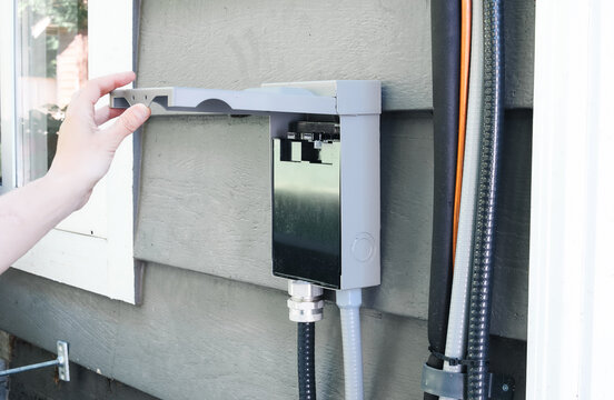 Electrician inspecting open AC disconnect box mounted above heat pump. Close up of electrical safety switch with conduit, wiring and refrigerant lines on exterior wall. Selective focus.