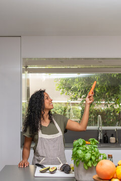 Smiling Hispanic woman holding carrot with copyspace in modern kitchen