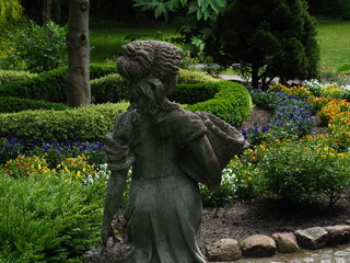 Fototapeta premium Back view of a classic woman statue holding a basket looking at the lush green botanical garden landscape - Vintage bronze sculpture standing in a German botanischer garten surrounded by colorful flow
