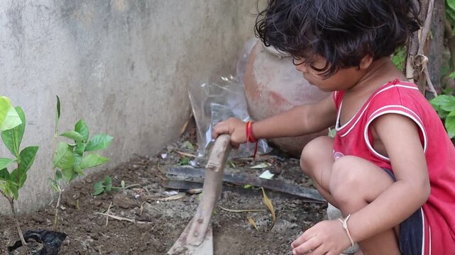Candid shot of a curious little Indian toddler girl actively engaging in gardening, using a hand tool to dig soil next to a green sapling, highlighting early childhood development and activity-based l