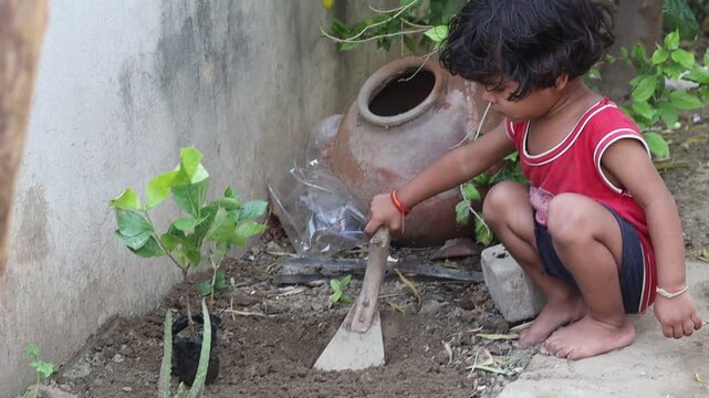 Candid shot of a curious little Indian toddler girl actively engaging in gardening, using a hand tool to dig soil next to a green sapling, highlighting early childhood development and activity-based l