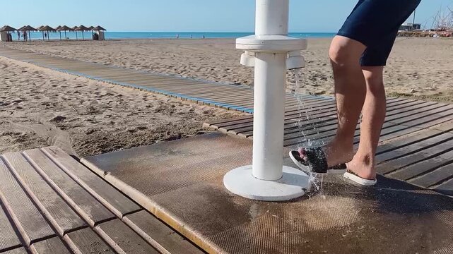 Close up of a man's feet wearing flip-flops while rinsing off at a public beach foot wash on a wooden boardwalk, with the sea in the background, representing beach hygiene, summer vacations, coastal l
