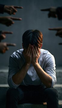 Stressed adult sitting on steps covering face while multiple hands point in accusation