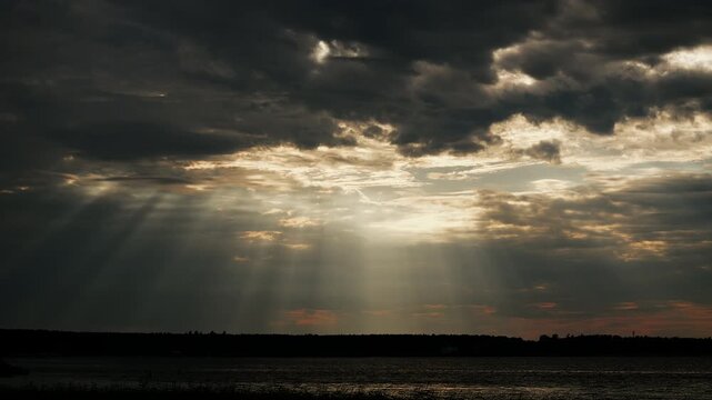 Powerful sun rays breaking through dramatic storm clouds above a calm lake. Atmospheric landscape with moody sky, crepuscular rays and changing weather.