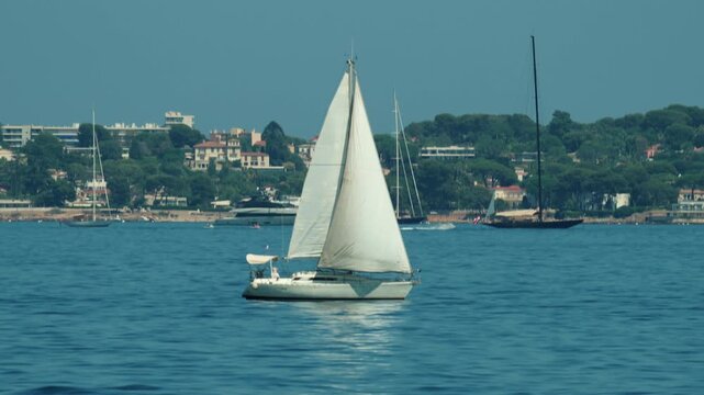 White sailboat cruising on blue calm water with a lush green coastal town in the background under a clear sky, Golfe Juan, France