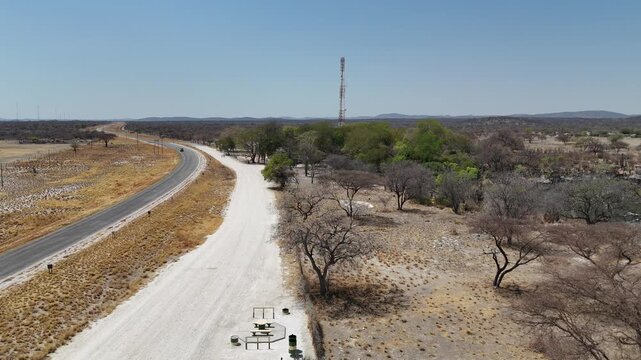 Cinematic aerial footage of a paved highway passing a small green oasis and a tall telecommunications tower in the dry Namibian savanna.
