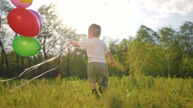 Boy running fast across field with balloon. Toddler smiles while holding balloon string. Summer grass around child. Balloon bounces in air. Outdoor sunlight shines on field, toddler, and balloon scene