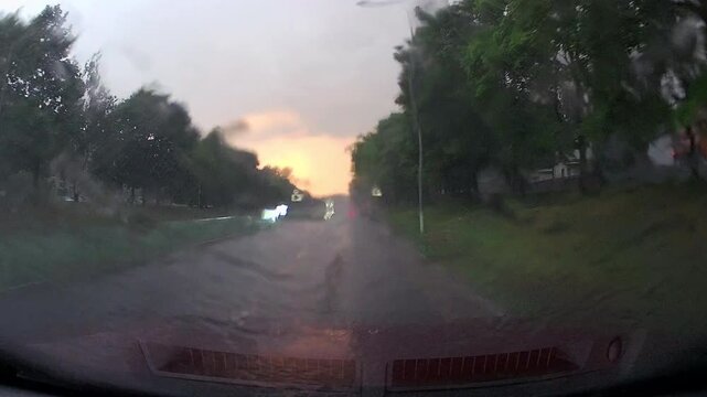 Dash Cam Video Of Rainy Road In City
Cars drive down a city road during rainy weather. The sunset creates a glow in the sky. Trees line the street. Water collects on the pavement. Its pouring. 