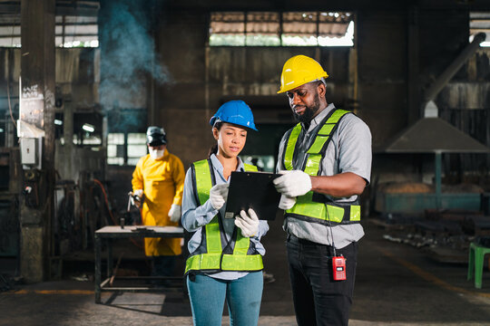 Worker factory helmet safety vest tablet inspection industry, teamwork with protective gear in metal workshop, focused employee checking digital report during production task