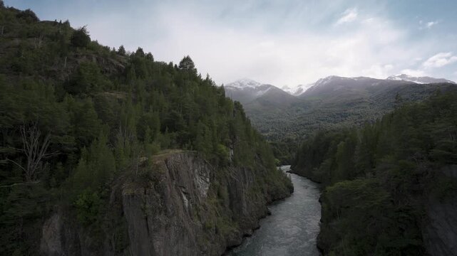 River flowing through a deep forest canyon, surrounded by lush trees, rocky cliffs, and distant snow-capped mountains.