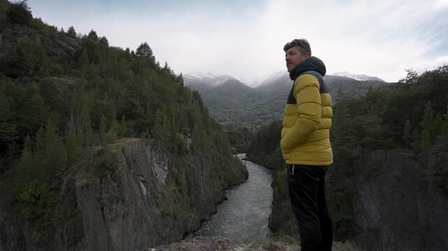 A man in a yellow and black jacket standing by a tree, observing the Futaleufu River flowing through a deep forest canyon in Patagonia, Chile.