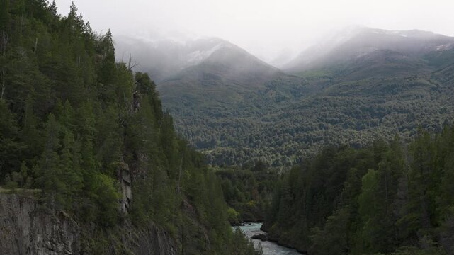 View of a deep forest canyon featuring lush trees and rocky cliffs, with snow-capped mountains and mist in the background, in Futaleufu, Patagonia, Chile.