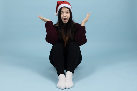 Confused young woman in santa hat shrugs shoulders in studio