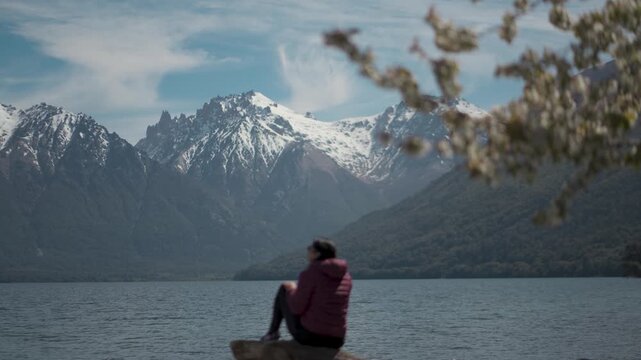 Woman sitting on a tree stump by Mascardi Lake, admiring the scenic view of snow-capped mountains and blooming cherry blossom branches in Patagonia, Argentina.