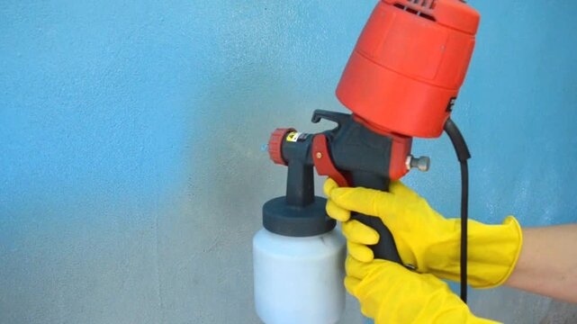 Worker wearing yellow gloves holds an electric spray gun and paints a vertical plaster surface. The process of applying a layer of blue paint to a wall using a hand-held spray gun. Renovation