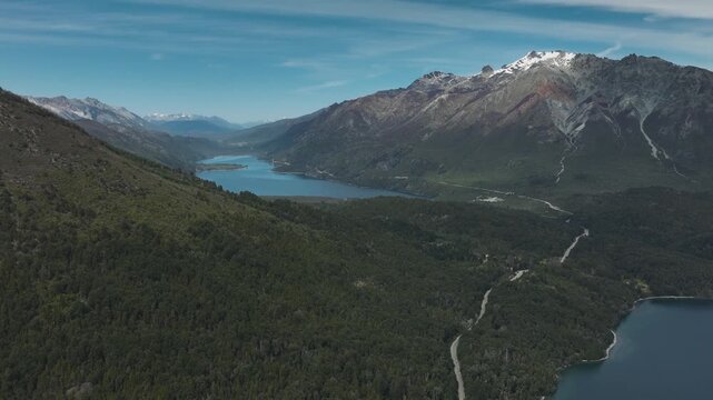 Aerial drone view of a vast mountain range with snow-capped peaks and lush forest valley surrounding a deep blue lake in Patagonia