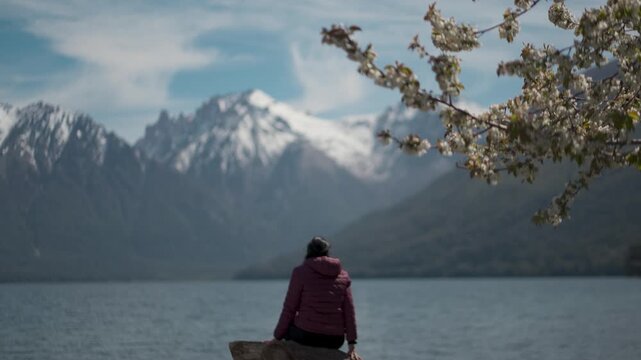 Woman looking at snow-capped mountains and a lake, framed by blooming white cherry blossom branches in a scenic spring landscape in Patagonia