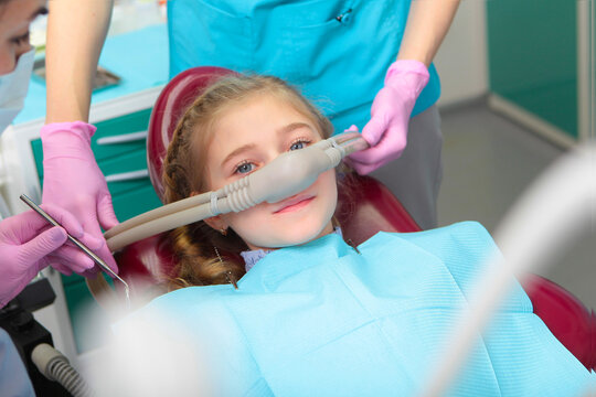 A little girl is comfortable to treat her teeth under superficial sedation. The girl smiles and holds two thumbs up. Milk teeth treatment. Treatment of children's teeth with nitrous oxide.