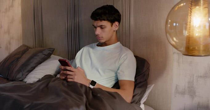 Adult male holding red-cased smartphone wearing smartwatch, tapping and scrolling, smiling in bed