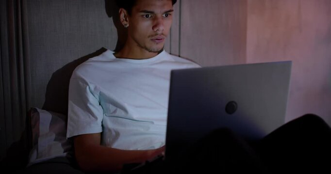 African American man sitting by headboard with laptop on lap in plain tee, reacting to screen