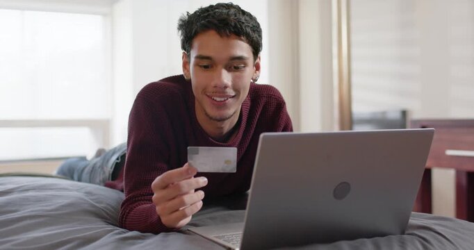 African American man propping on elbows holding credit card studying laptop in bedroom buying