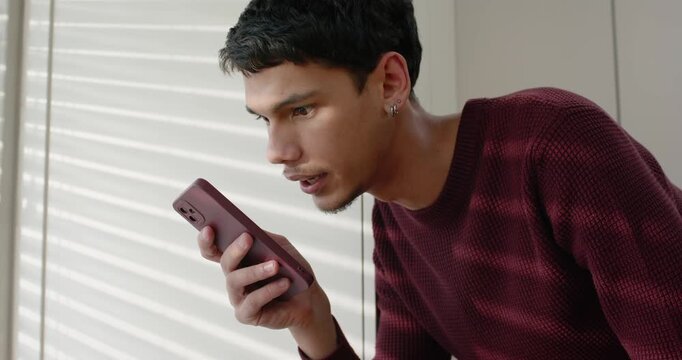 Man in sweater holding pink phone near mouth by blinds, speaking, typing on white laptop, glancing