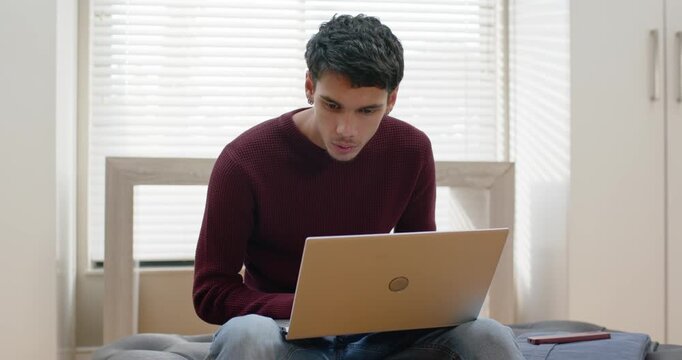African American 20s male sitting on bed, working on laptop beside smartphone leaning and typing
