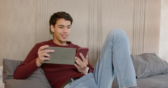 African American man in maroon sweater reacting to phone alert while tapping folio tablet on bed