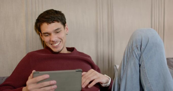 African American man reclining on sofa glancing at phone then reading tablet content and laughing
