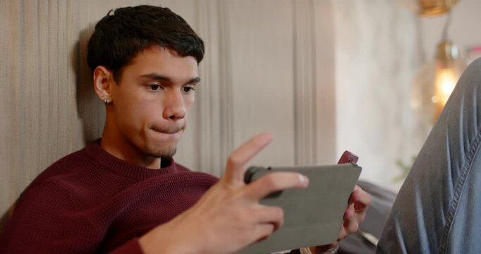 African American man reclining on sofa holding tablet, tapping cover and reacting to content