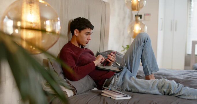 Man in twenties reclining on bed, tapping tablet then picking up smartphone, responding to messages