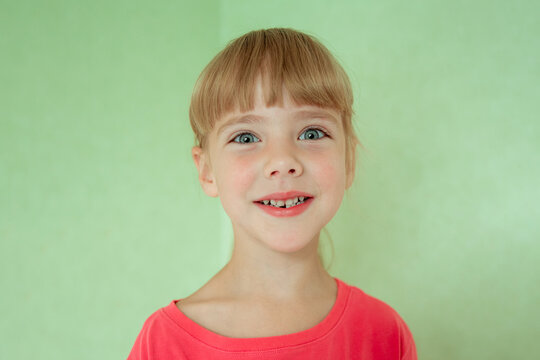 Young girl with a bright smile showing a gap from missing front teeth, blonde hair with bangs, wearing a coral pink top, soft pastel floral background